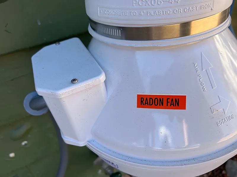 Close-up of a radon fan housing showing its RADON FAN label and moulded air-flow arrow