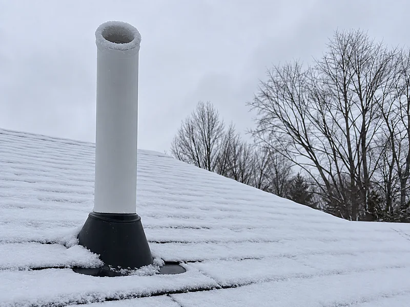 A radon vent stack discharging above a snow-covered roof, its rim edged with frost
